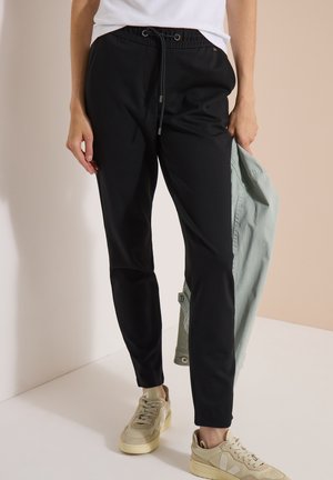 Person trägt schwarze Kordelhose, weiße Sneakers und hält eine hellgrüne Jacke vor einem beigen Wandhintergrund.