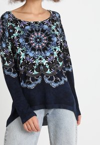 Donna che indossa un maglione navy a maniche lunghe con un intricato motivo a mandala blu, viola e rosa, abbinato a jeans azzurri chiari.