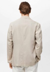 Chaqueta beige claro con un corte relajado, que cuenta con mangas largas, una abertura trasera única y detalles de costura visibles. Textura suave.