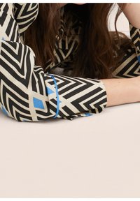 Tela de patrón negro y crema con acentos azules, que presenta un diseño geométrico. La prenda tiene mangas enrolladas que exhiben el patrón.