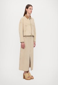 Giacca beige corta con colletto marrone, ampia tasca frontale, abbinata a una lunga gonna beige con uno spacco frontale. Indossata con zoccoli con suola in legno chunky.