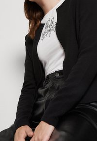 Czarny dziergany kardigan z teksturowaną powierzchnią, noszony na białej koszuli z czarnym nadrukiem graficznym. Model siedzący, ubrany w spodnie z imitacji skóry.