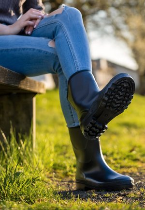 Bottes en caoutchouc noires avec des semelles texturées et une finition lisse, posées dans l'herbe à côté d'un jean bleu avec des genoux usés sur un banc en bois.