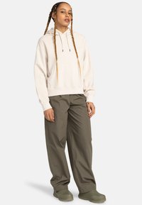 Beige hooded sweatshirt with a kangaroo pocket, paired with loose olive green pants and beige slip-on shoes. Long braided hair.