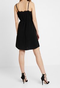 Schwarzes ärmelloses Kleid mit Spitzenbesatz am Ausschnitt, tailliert und mit einem fließenden Rock. Kombiniert mit schwarzen, hochhackigen Sandalen mit Riemchen.