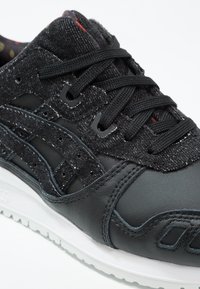 Svart träningsskor med ovandel i läder och strukturerad textil, försedda med perforeringar, svarta snören och en vit gummisula.