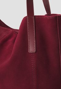 Gros plan d’un sac cabas en daim rouge profond montrant une anse en cuir lisse cousue au corps du sac.