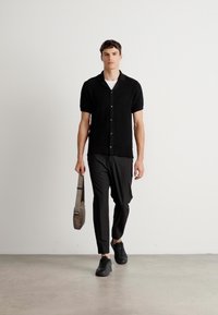 Man loopt binnen en draagt een zwart, kortemouw, knoop shirt, zwarte broek, zwarte sneakers en houdt een grijze schoudertas in zijn linkerv hand.
