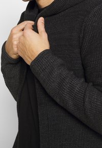 Cardigan scuro con motivo a coste e collo alto, parzialmente chiuso davanti, indossato sopra una maglia nera.