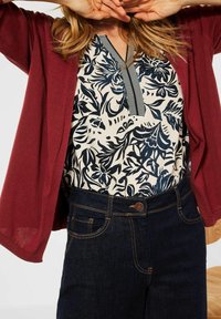 Cárdigan burdeos sobre una camisa floral en tonos navy y crema con un cuello de rayas vertical, combinado con unos jeans de denim oscuro.
