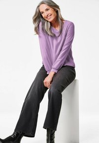 Lila Langarmoberteil mit gerafften Manschetten, kombiniert mit dunklen Jeans. Das Modell trägt schwarze Stiefeletten und sitzt auf einem weißen Podest.