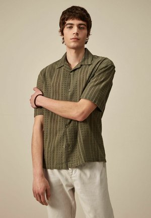 Camicia a maniche corte di colore verde oliva, caratterizzata da un tessuto a motivo testurizzato con bottoni e colletto, abbinata a pantaloni di colore chiaro.