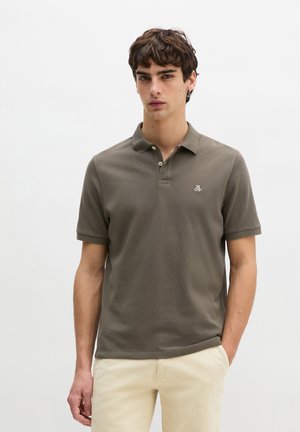 Młody mężczyzna w oliwkowozielonym polo z krótkim rękawem i beżowych spodniach, stoi na tle jednolitego białego tła.