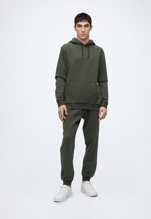 Ung mand står iført en mørkegrøn hættetrøje og matchende joggebukser med hvide sneakers mod en ensfarvet lys baggrund.
