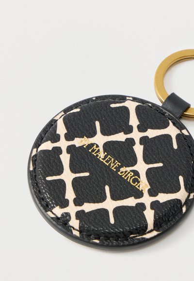 Στρογγυλό μπρελόκ με σχέδιο σε μαύρο και μπεζ χρώμα, με χρυσό κρίκο και τυπωμένο το "DI MALENE BIRGER" σε χρυσά γράμματα.