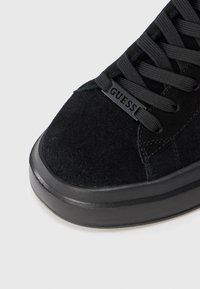 Melna zamša sporta apavi ar melnām auklām un metāla birku ar zīmola nosaukumu "GUESS" priekšā.