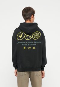 Sweat à capuche noir avec un grand motif jaune au dos, comprenant des icônes circulaires et du texte. Confectionné dans un tissu doux, il présente une coupe décontractée et une capuche.