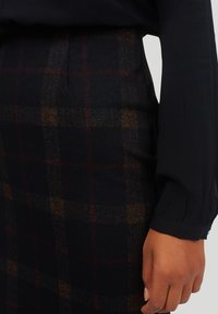 Blandad ullpennkjol med ett mörkt rutmönster i röd och brun, tillsammans med en långärmad svart blus.