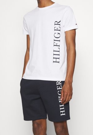 T-shirt en coton blanc avec "HILFIGER" imprimé verticalement en noir, associé à un short en jersey bleu marine présentant le même logo en blanc.