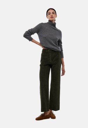 Pull en cachemire gris, pantalon en velours côtelé vert à jambes larges, chaussures en daim marron. Le mannequin se tient avec assurance, les mains sur les hanches, contre un fond blanc.