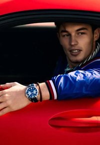 Orologio blu con caratteristiche cronografiche al polso, indossato con un bracciale di pelle marrone. Braccio appoggiato su una porta di auto rossa, indossando una giacca blu.