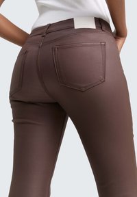 Pogled od blizu na osebo, ki nosi prilegajoče rjave hlače z zadnjimi žepi in belo srajco, zataknjeno v hlače, ki stoji proti enobarvni svetli ozadju.