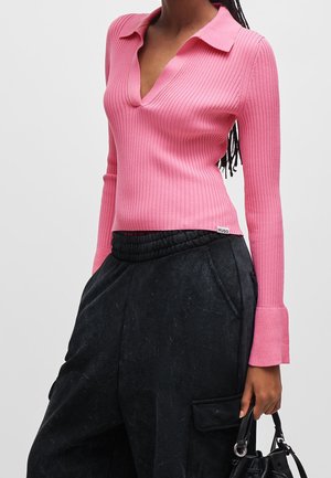 Vrouw draagt een roze geribd lang mouw top met kraag, zwarte wijde broek, houdt een zwarte handtas bij zich.