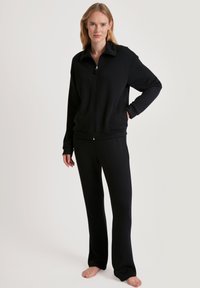 Giacca nera con zip e colletto alto, abbinata a pantaloni neri, entrambi realizzati in tessuto morbido. Vestibilità rilassata con maniche lunghe e polsini.