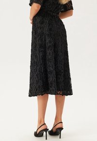 Schwarzes Spitzenkleid mit floralem Muster, knielang, strukturierter Stoff, kurze Ärmel, tailliert, kombiniert mit schwarzen High Heels.