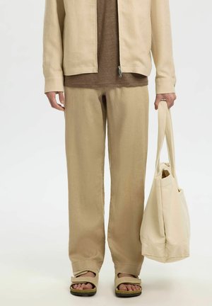 Personne portant un pantalon beige, une veste beige sur une chemise marron, des sandales beiges, tenant un grand sac fourre-tout beige sur le côté.