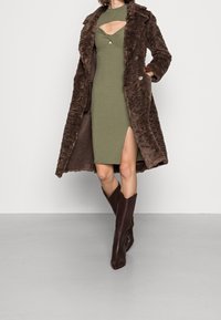 Manteau en fausse fourrure marron texturée porté sur une robe côtelée vert olive avec un design découpé et une fente latérale, associé à des bottes marron à bouts pointus.