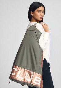 Zielona, lekka kurtka z błyszczącym brązowym obszyciem z napisem "CLUB" w białym kolorze. Prosty design, widoczne przeszycia i ściągacz u dołu.