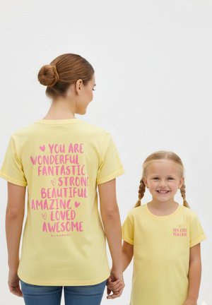 Woman and girl wearing matching yellow shirts with pink positive affirmations, holding hands; woman faces away, girl smiles at camera.