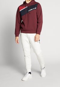 Sudadera marrón con un diseño diagonal en azul marino y rojo, con texto en blanco. Combinada con pantalones de chándal gris claro y zapatillas blancas.
