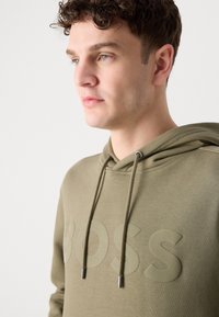 Felpa verde oliva con logo "BOSS" testurizzato. Presenta un cappuccio con cordoncino e lacci con estremità metalliche. Materiale morbido con una vestibilità casual.