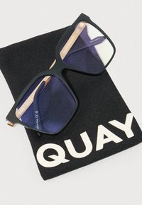 Čierne slnečné okuliare s modrými šošovkami, kovovými detailmi a matným povrchom, vystavené na čiernom textilnom vrecku s nápisom "QUAY."