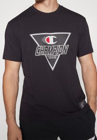 Musta puuvillapaita, jossa on iso kolmiomainen graafinen kuva, jossa on valkoinen ja punainen "CHAMPION"-teksti ja logo. Paidassa on lyhyet hihat ja pyöreä kaula-aukko.