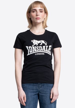 Žena s rovnými svetlými vlasmi nosí čierne tričko Lonsdale London a modré džínsy, otočená vpred na jednoliatom bielom pozadí.