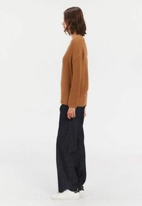 Un pull en tricot brun texturé avec un ourlet côtelé, associé à un pantalon en denim sombre ample et des baskets blanches, vu de côté.