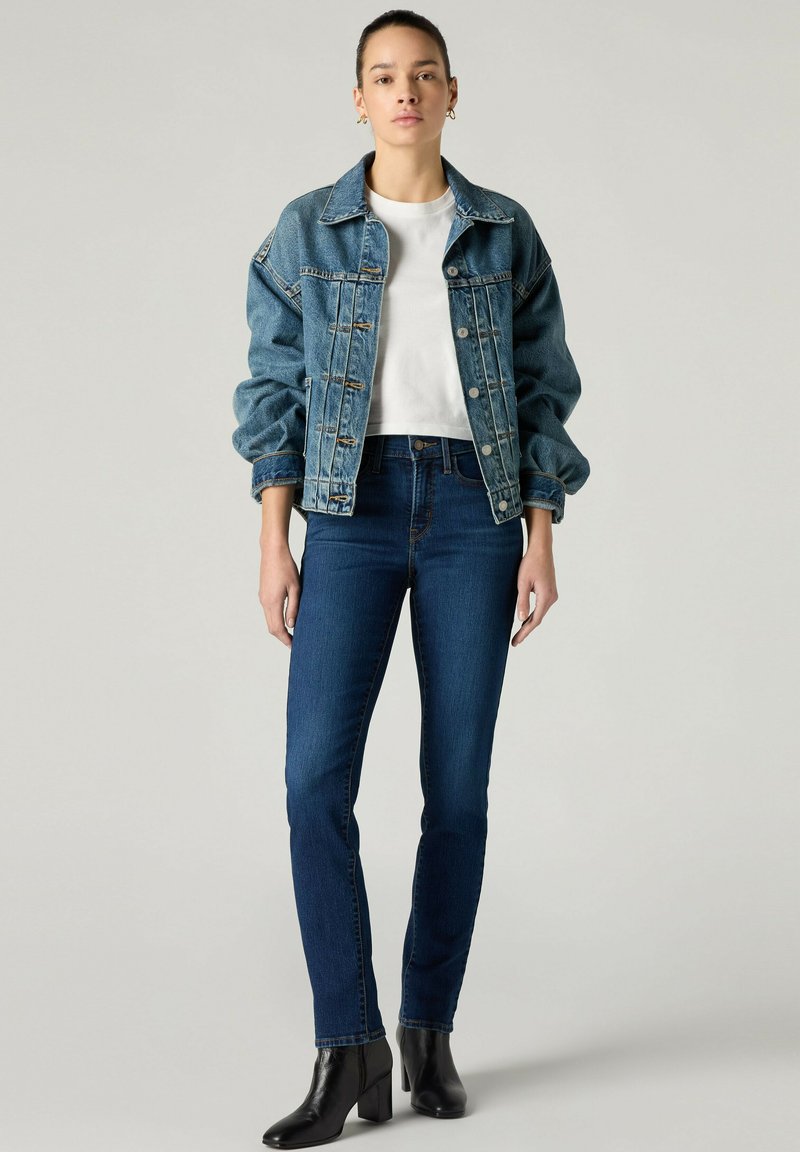 Jeune femme portant un t-shirt blanc, une veste en jean bleu oversize, un jean skinny bleu foncé et des bottines noires, debout devant un fond uni.