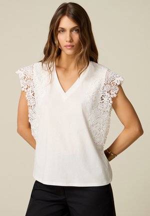 Woman wearing a white sleeveless blouse with floral lace details on the shoulders and black pants against a plain background.
