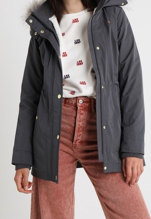 Femme portant une veste à capuche gris foncé sur une chemise blanche imprimée et un pantalon en velours côtelé couleur rouille taille haute, debout les mains détendues.