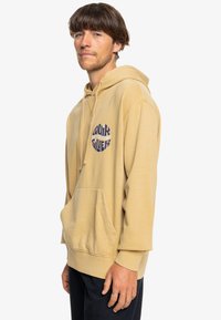 Beige hooded sweatshirt made of soft fabric, featuring a kangaroo pocket and a circular navy logo design on the chest. Relaxed fit.