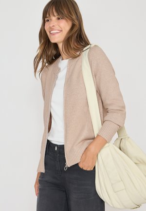Beige Strickjacke mit geripptem Saum und Bündchen, getragen über einem weißen Hemd. Kombiniert mit dunklen Jeans und einer hellen, lässigen Handtasche.