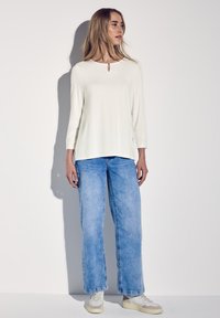 Top blanco de manga larga con un escote en forma de lágrima, combinado con pantalones vaqueros de pierna ancha de color azul claro y zapatos blancos. Tejido suave, ajuste casual.
