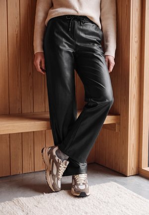 Personne posant devant un mur en bois, vêtue d’un pantalon en cuir noir, d’un pull en maille beige et de baskets couleur taupe.