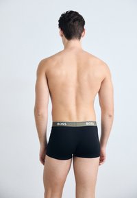 Schwarze Boxer-Briefs mit einem grün-weißen "BOSS"-Logo auf dem Bund. Glatte Stoffoberfläche mit engem Sitz, von hinten betrachtet.