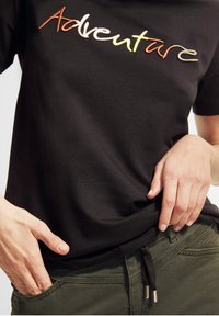 Camiseta negra de algodón con texto bordado colorido que dice "Aventura", combinada con pantalones verdes con cordón y detalles de hardware en tono plateado.