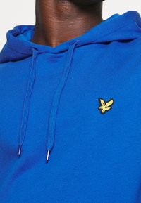 Primo piano di una persona che indossa un hoodie blu con un'emblema di un uccello giallo sul petto e lacci blu.