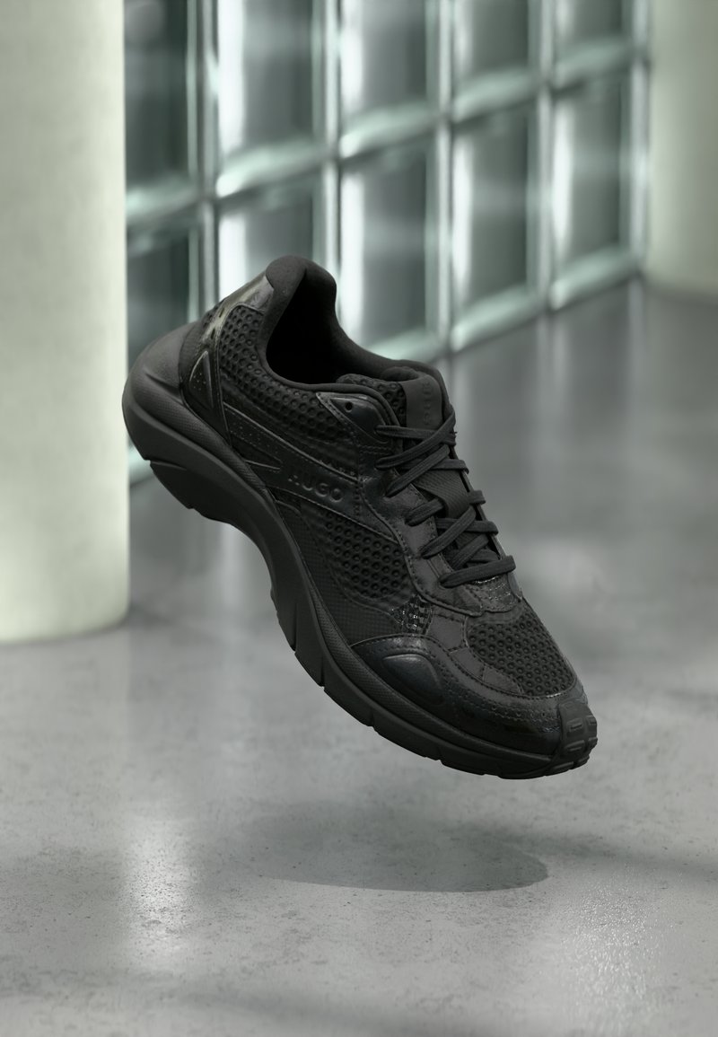 Baskets athlétiques noires avec détails en mesh et cuir, flottant au-dessus d'un sol en béton poli près d'un mur en blocs de verre dans un espace intérieur minimaliste.